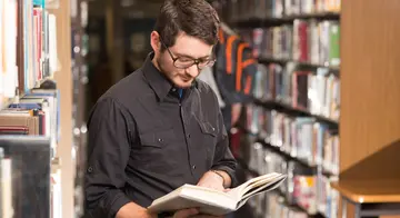A man is standing and reading a book with library shelves full of book behind him.