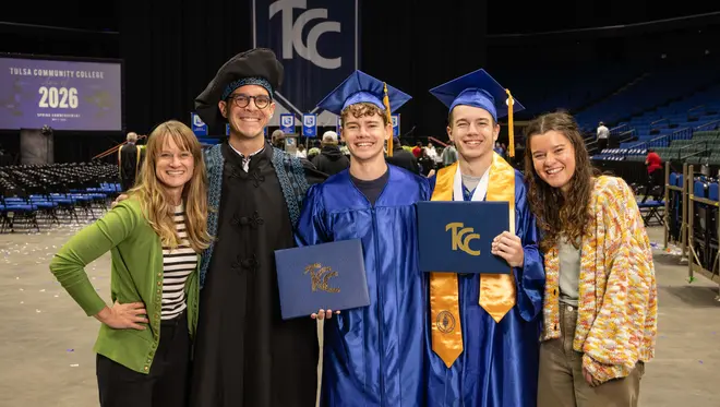 Ashley, Professor Da Corte, Hudson, Everett, and Breese pose for a photo and smile. Hudson and Everett are wearing blue graduation regalia and hold their degrees.