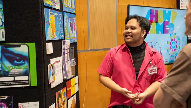 Digital Media student Keith Ponce speaks with spectators about his work. He is looking at his portfolio which is displayed on a wall.