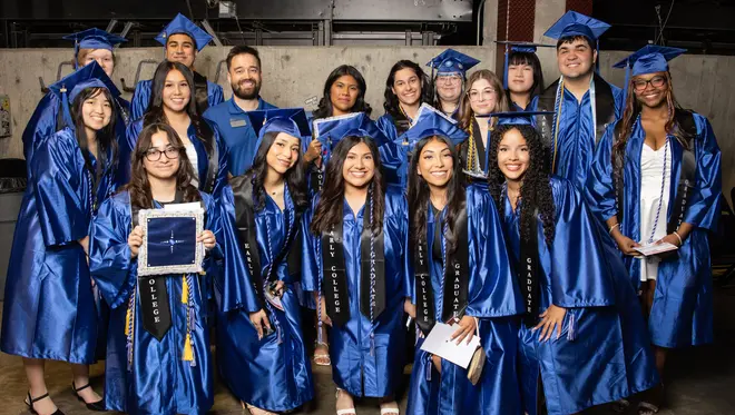 A group of 16 concurrent graduates smile in their regalia before TCC's Spring 2026 Commencement Ceremony. A TCC Dual Credit employee smiles with them.