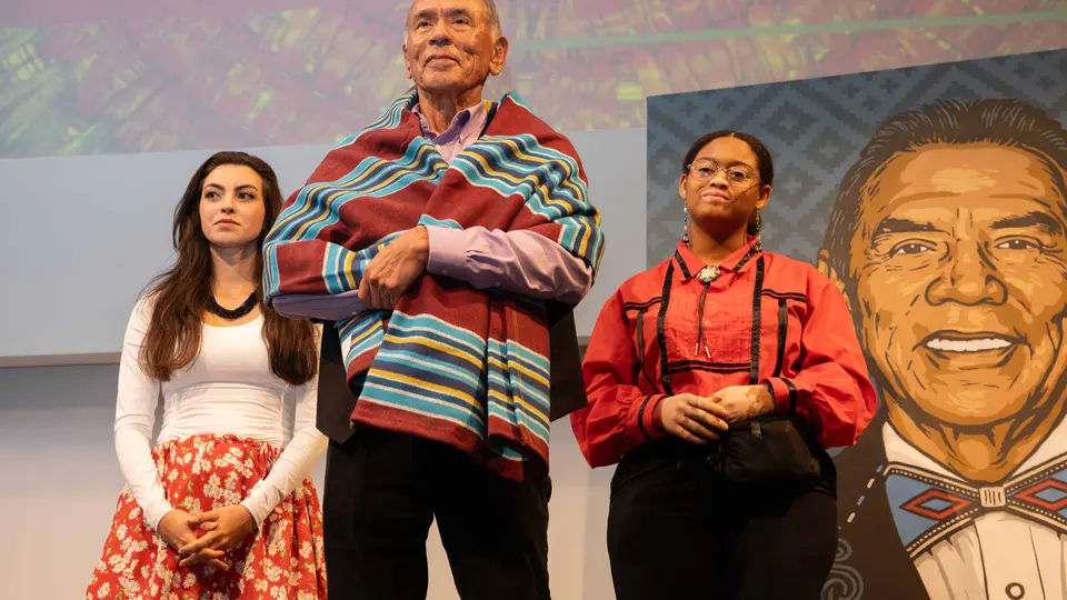 A TCC students stands on either side of Wes Studi, who is wrapped in a blanket that has a Native American design.