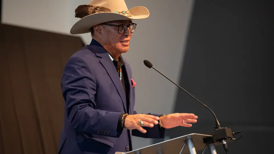 Mark A. Wilson-Fourkiller is standing at a podium and speaking. We is wearing a western-style hat with a feather in it and a blue suit jacket.