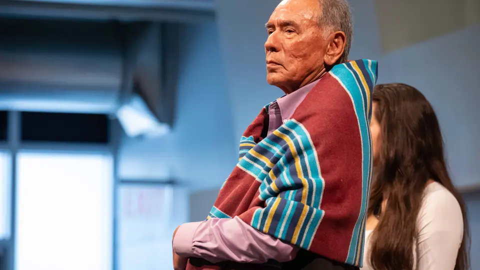 Wes Studi is standing and wrapped in a blanket. The blanket has a Native American design and bright colors of red and blue.