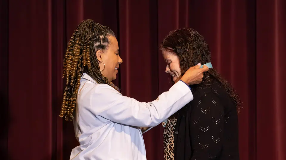 Faculty Dr. Angela Elmore drapes a ribbon with a Nursing pin around a student's neck. The student is a woman wearing a leopard print dress and black sweater. Both people are smiling.