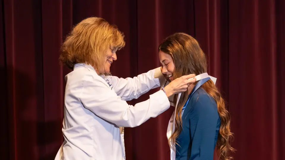 Faculty Susan Goldsmith drapes a ribbon with a Nursing pin around a student's neck. The student is a woman wearing a blue dress. Both people are smiling.