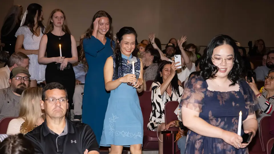 Students each holding one white candle walk down stairs in the VanTrease PACE to be seated for the ceremony. A woman in a blue dress is smiling.